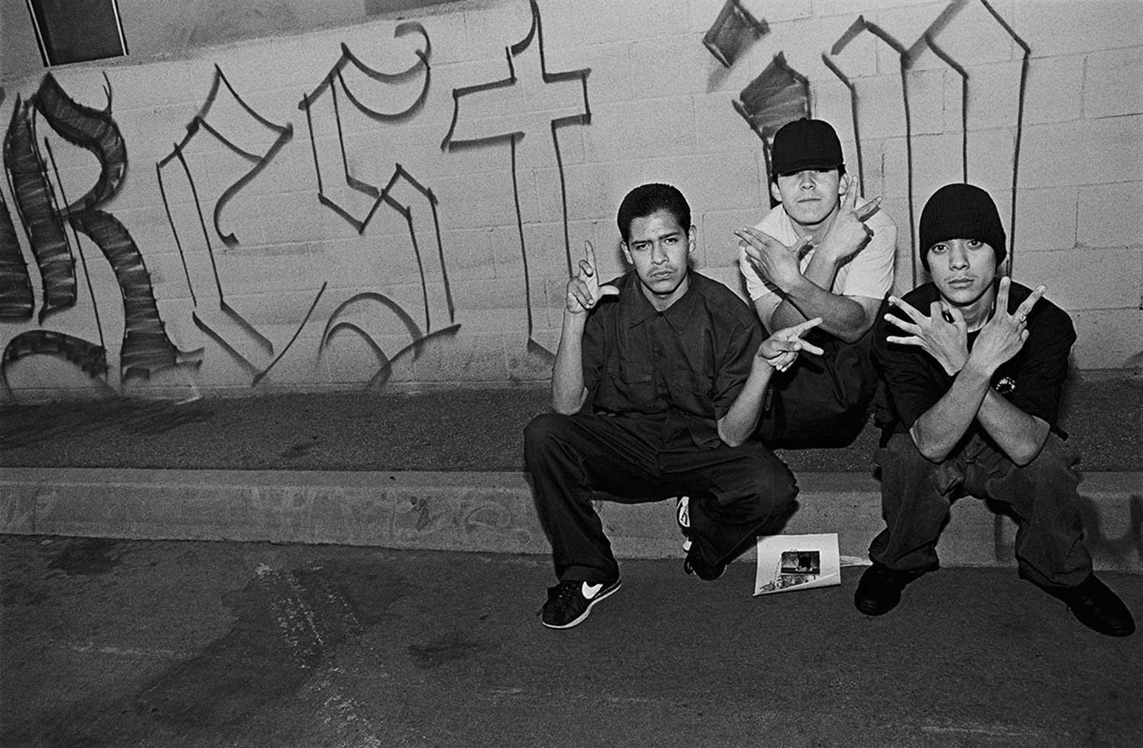 Thre Latino gang members throwing hand signs in front of RIP mural. 