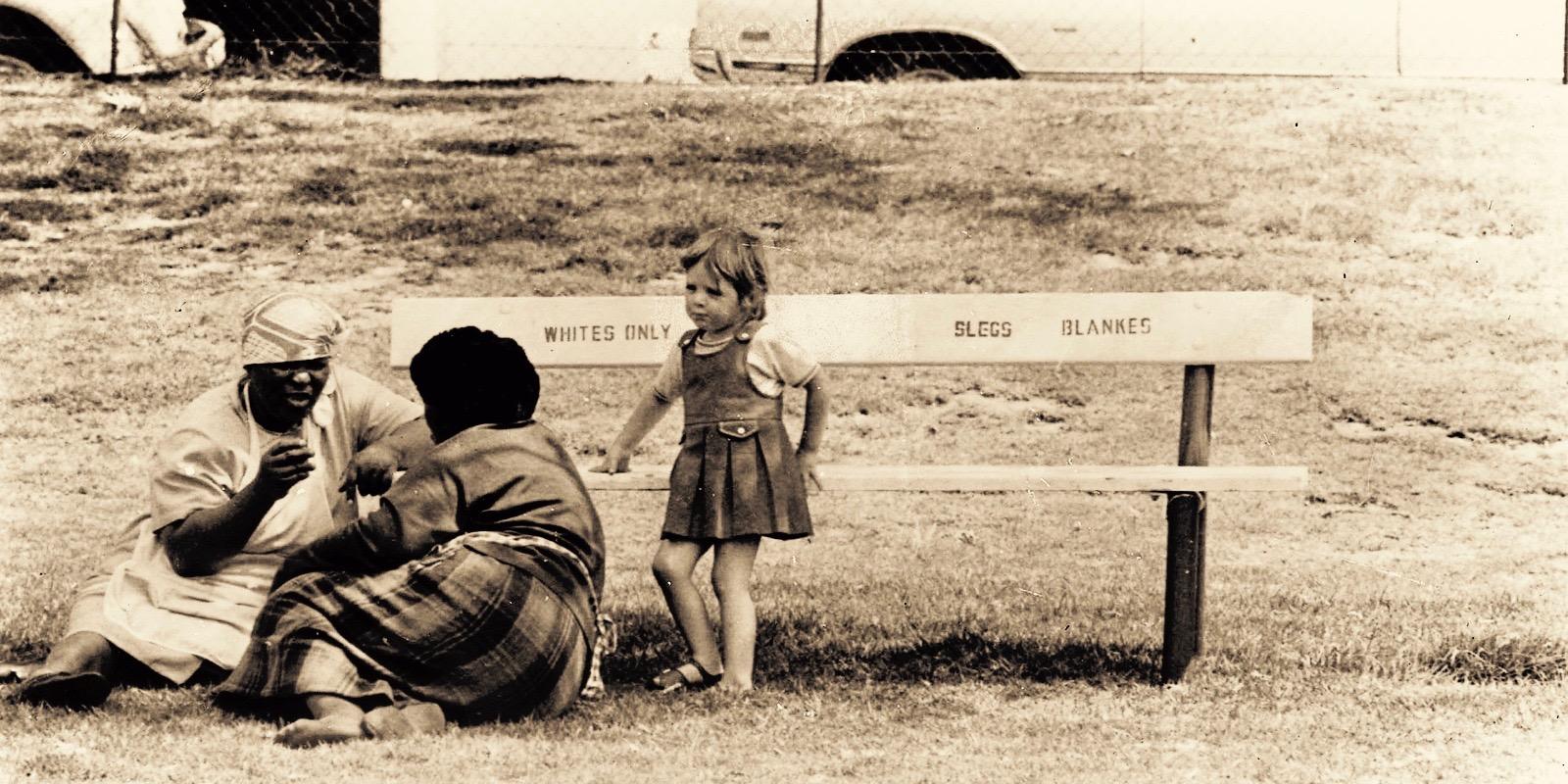 white girl child near a bench for whites only, with two black women sitting on the grass